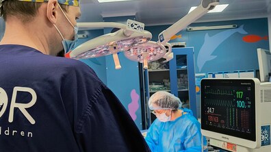 Фото з операційної, де двоє медиків процедуру. У центрі фотографії операційний світильник, а поруч із ним медики в масці, шапочці та халаті. Ліворуч стоїть монітор, на якому відображається серцебиття та інші важливі показники пацієнта.