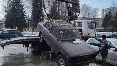 На вулиці кран евакуатора підіймає темно-фіолетовий легковий автомобіль з багажником на даху, працівник поруч координує процес. Навколо видно припарковані авто, дерева та будинки