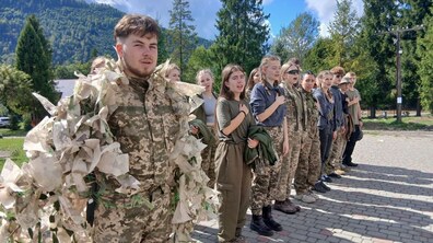 Учасники військово-патріотичного вишколу, вдягнуті у військову форму, співають гімн України. Позаду видно гори