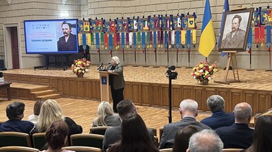 Заступниця міського голови Львова Ірина Кулинич виступає за трибуною в актовій залі ЛНУ ім. Івана Франка під час відкриття Міжнародного наукового конгресу. На другому плані видно ряд хоругов факультетів університету, прапор України та фрагмент прапора ЄС, портрет Івана Франка на мольберті та великий екран із презентацією події.