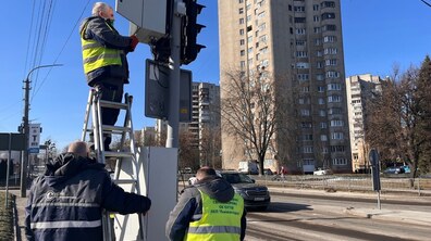 Троє працівників ЛКП «Львівавтодор» виконують монтажні роботи: чоловік у темному спецодязі з яскравим жовтим світловідбивним жилетом стоїть на драбині та встановлює блок живлення на опорі світлофора, унизу допомагають двоє колег — один також у жилеті, інший у темній куртці без світловідбивних елементів. На тлі видно дорогу з автомобілем, дерева без листя, багатоповерхові будинки та блакитне небо.