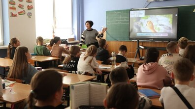 У класі вчителька стоїть біля дошки та інтерактивного екрана перед учнями, які сидять за партами