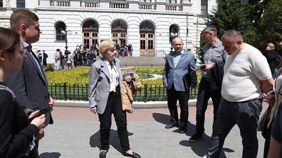 Група людей стоїть перед головним корпусом Львівської політехніки. У центрі — Ірина Маруняк, у світло-сірому піджаку та чорних штанах, яка спілкується з учасниками зустрічі просто на площі перед будівлею. Навколо неї — кілька чоловіків у діловому одязі, один із них тримає папери та щось пояснює. На задньому плані видно світлий фасад історичної будівлі з арковими дверима та клумбами перед входом.