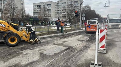 Ділянка міської дороги під час підготовчих робіт до ремонту покриття. На передньому плані видно зрізаний асфальт смугами, поверхня нерівна, з темними смугами та заглибленнями — це підготовлена основа перед укладанням нового шару. Праворуч стоять тимчасові дорожні щити з червоно-білими діагональними смугами, які відмежовують робочу зону від транспорту. На тротуарі поруч із дорогою стоять двоє працівників у помаранчевих сигнальних жилетах.