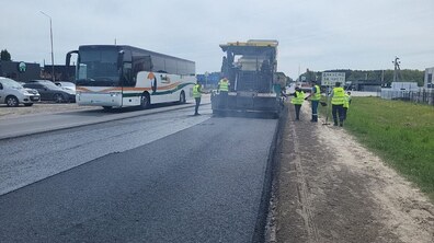Дорожники у світловідбивних жилетах ремонтують одну зі смуг дороги: працівник вкладає нове темне покриття, поруч стоїть дорожня техніка та працівники з лопатами, а сусідньою смугою повільно проїжджає великий білий автобус, навколо — узбіччя з травою, кілька автомобілів і похмуре сіре небо.