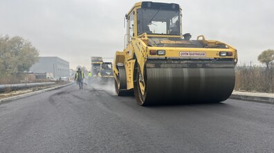 На свіжому асфальті працює спецтехніка, що ущільнює покриття. Позаду видно кількох працівників у сигнальних жилетах. Вздовж дороги облаштовані бордюри, а на задньому плані розташовані промислові будівлі й дерева.