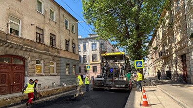 Ремонт дороги на вул. Героїв УПА. У центрі працює асфальтоукладальна машина, поруч кілька дорожніх робітників у жовтих світловідбивних жилетах укладають свіжий чорний асфальт. Обабіч стоять старі багатоповерхові будинки, а праворуч височіє велике зелене дерево, що затіняє частину вулиці.