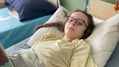 11-річна Оля лежить на лікарняному ліжку. На обличчі дівчинки окуляри з темною оправою. Оля одягнена у світлу жовту футболку, накрита ковдрою зі світлим візерунком.