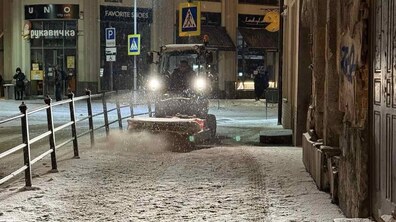 Нічна міська вулиця під час легкого снігопаду. Праворуч — стіна старої будівлі з дверима, ліворуч — металевий поручень, що відділяє тротуар від проїжджої частини. Посеред тротуару їде маленький тракторець. Попереду трактора встановлена щітка або відвал — він активно змітає тонкий шар снігу з плитки. Удалині стоять кілька людей, а на будівлях видно вивіски магазинів і кафе.