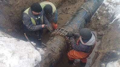 Троє працівників Львівводоканалу працюють у глибокій земляній траншеї. У центрі кадру – велика чорна пластикова труба водопроводу, розташована горизонтально. Її з’єднують спеціальною металевою муфтою з болтами. Один із чоловіків тримає інструмент і закручує болти на з’єднанні, інші допомагають притримувати та фіксувати конструкцію.