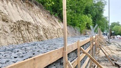 будівництво підпірної стіни для укріплення схилу біля дороги на в’їзді у Львів. По центру тягнеться довга конструкція з габіонів — металевих сіток, заповнених сірим камінням. Вона укріплена тимчасовим каркасом із дерев'яних дошок.