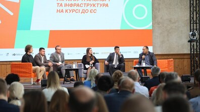 Головна сцена IV Urban Mobility Forum. На підвищенні сидять п’ятеро чоловіків та одна жінка, а один із учасників говорить у мікрофон. Позаду них встановлено великий яскравий екран із написом помаранчевими літерами: «ЄВРОПЕЙСЬКИЙ ШЛЯХ УКРАЇНИ: ТРАНСПОРТ ТА ІНФРАСТРУКТУРА НА КУРСІ ДО ЄС». На передньому плані видно силуети людей в аудиторії, які слухають виступ.