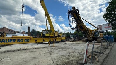 Будівельний майданчик із великою жовтою спеціалізованою технікою для буріння та влаштування фундаментів. У центрі працює кран із підвішеним тросом, праворуч стоїть потужна бурова установка на гусеницях. Навколо видно бетонний майданчик, допоміжну техніку, огорожі та мобільні туалети. На тлі — дерева, будівлі та хмарне небо з блакитними просвітами