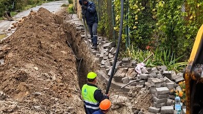 Троє працівників виконують ремонтні роботи на вузькій вулиці. Вони працюють у глибокій траншеї, уздовж якої розсипана вийнята земля. Один із працівників стоїть біля краю траншеї, двоє інших перебувають усередині. Обабіч вулиці ростуть дерева та кущі, а на задньому плані видно приватні будинки.