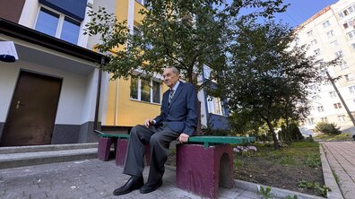 Мешканець будинку на вул. Гната Хоткевича, 56 сидить на лавці у дворі біля оновленого фасаду будівлі. Позаду чоловіка видно дерева, квіти та прибудинкову територію