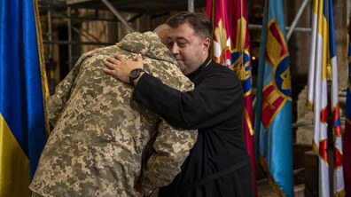 Настоятель Гарнізонного храму святих апостолів Петра і Павла – отець Тарас Михальчук обіймає військовослужбовця, який повернутий до камери спиною. На фоні є великі прапори Збройних Сил України, які розміщені у храмі.