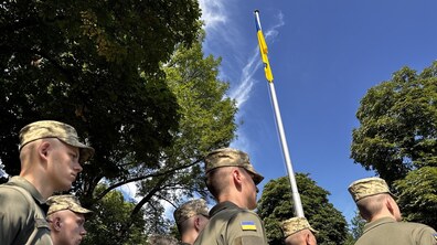 Курсанти стоять на тлі зелених дерев під час урочистого підняття державного синьо-жовтого прапора на високому флагштоку