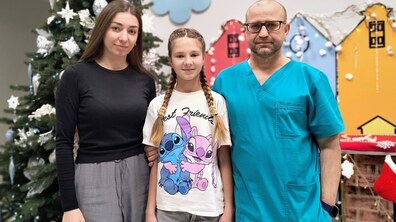 У холі дитячої лікарні святого Миколая стоять: усміхнена пацієнтка Дарина, її мама та хірург Олександр Калінчук. На задньому плані – прикрашена ялинка з білими сніжинками та іграшками, декоративний камін із різдвяною шкарпеткою та кольорові декоративні будиночки на стіні