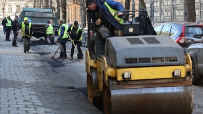 На міській вулиці комунальники ліквідовують ями гарячим асфальтом. На передньому плані дорожній каток ущільнює свіже покриття, а позаду працівники у світловідбивних жилетах розрівнюють асфальт лопатами біля вантажівки. Обабіч дороги видно брукований тротуар, припарковані автомобілі та фасади старих будинків.