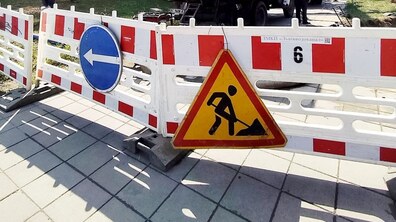 Фото ілюстративне. На вулиці стоїть біло-червона огорожа, на якій розміщені дорожні знаки: синій зі стрілкою вліво та жовтий трикутний «Дорожні роботи». За огорожею на другому плані стоїть біла вантажівка, також видно працівників.