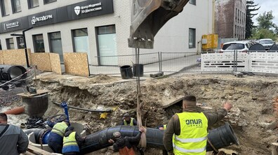Працівники у світловідбивних жилетах прокладають нову водопровідну трубу у котловані, використовуючи екскаватор. На другому плані видно будівлю та автівки