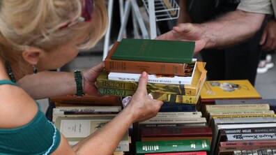 жінка тримає в руках стос книжок, серед яких помітні видання із зеленими, жовтими, помаранчевими та білими обкладинками, а на столі перед нею розкладено ще багато книжок різних кольорів і форматів. Навколо видно книжковий стенд, де люди переглядають та обирають літературу.