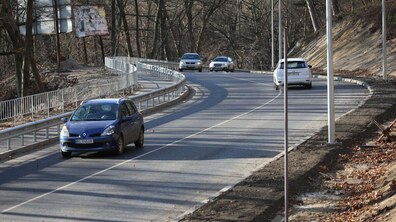Ділянка дороги на вул. Личаківська на виїзді з міста. Асфальтована вулиця плавно вигинається вправо і піднімається вгору серед лісистої місцевості. З лівого боку дороги — металеві відбійники, а праворуч видно схил із землею, сухим листям та новими електричними стовпами. На дорозі рухаються кілька автомобілів: найближче — синій легковик, далі ще кілька машин у обох напрямках.