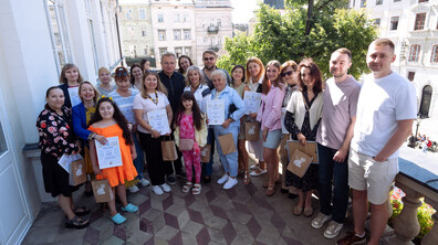 Переможці конкурсу, дорослі і діти стоять на балконі ратуші разом з міським головою і в руках тримають дипломи. Позаду них видніються сторвинні будівлі з шпилястими дахами