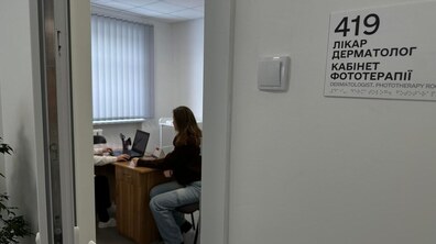 На передньому плані – вивіска «419, Лікар дерматолог, Кабінет фототерапії». У кабінеті видно жінку, яка сидить за столом, а поруч – медик.