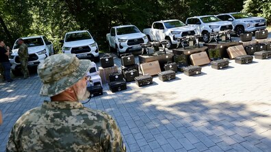 На відкритому майданчику стоять автомобілі, безпілотники та різне технічне обладнання, підготовлене для передачі військовим. Позаду видно дерева, а на передньому плані – військовослужбовець у формі й панамі, який стоїть спиною до камери.