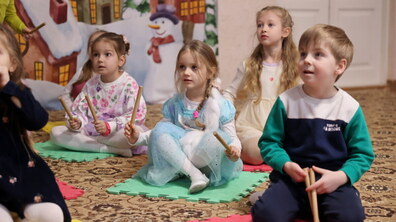 Діти дошкільного віку під час музичного заняття у дитячому садку. Малюки сидять на кольорових килимках із дерев’яними паличками в руках та уважно слухають вихователя.