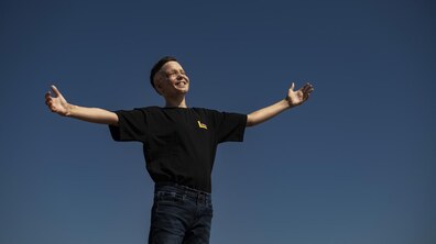 Львів’янин Ромчик Олексів стоїть на тлі яскравого блакитного неба. Він широко розвів руки в сторони та усміхається, заплющивши очі. На ньому – чорна футболка й темні джинси.
