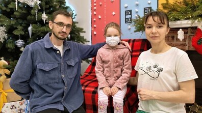Тато Сергій разом з донькою Дарією та дружиною Юлією сидять у холі дитячої лікарні святого Миколая, позаду – новорічна ялинка та святкові декорації