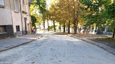 Відремонтована вул. Кубійовича - рівна брукована проїжджа частина. Ліворуч простягається оновлений тротуар вздовж житлових будинків, яким іде жінка у синій сукні. Вздовж дороги ростуть високі дерева.