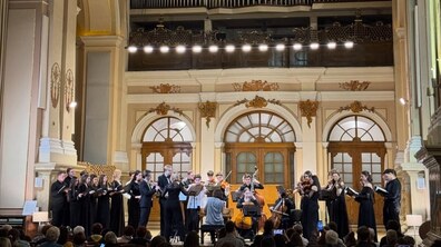 Концерт у розкішній залі з високими стелями та класичним декором у Львівському органному залі. На сцені виступає камерний оркестр і хор у чорному вбранні, а перед ними сидить диригент. У залі глядачі, які уважно слухають виступ і знімають на телефони.