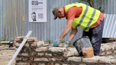 Робітник у жилетці мурує стіну зруйнованого музею Шухевича. На фоні позаду нього паркан, на якому плакат з написом “ Місто відновить музей” та портретом Романа Шухевича