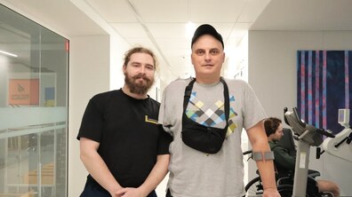 Пацієнт центру UNBROKEN Олег Трофімчук та його фізичний терапевт стоять у реабілітаційному залі й усміхаються. Обоє дивляться в камеру. Позаду видно, як на тренажері займається інший ветеран.