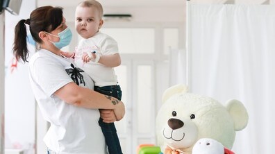 Медична працівниця тримає на руках маленьку пацієнтку Мирославу. Дитина дивиться прямо в камеру, а на її руці помітна пов’язка та медичний катетер. На передньому є великий білий плюшевий ведмедик та інші м’які іграшки.
