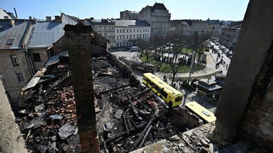 Зруйнований дах будівлі після російської атаки: обвуглені балки, уламки цегли та попіл. На задньому плані – міська площа з деревами, людьми та жовтим автобусом, що рухається вулицею. Контраст між знищеною будівлею на передньому плані та звичайним міським життям позаду підкреслює руйнівні наслідки російської агресії.