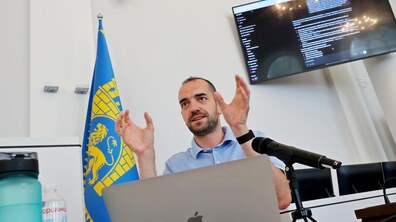 Спікер у світло-блакитній сорочці виступає перед учасниками тренінгу, сидячи за ноутбуком і пояснюючи матеріал на фоні великого екрану, на якому включена презентація