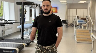 Захисник Руслан Сливка стоїть у реабілітаційному залі в чорній футболці та камуфляжних шортах. Він дивиться прямо в камеру. На задньому плані видно бігові доріжки, тренажери та спеціальне обладнання для фізичної реабілітації.