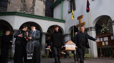 Люди несуть труну із тілом Олени Рибалко через подвір’я старовинного храму в центрі Львова. Поруч іде священник у церковному вбранні, а на задньому плані видно аркові галереї, кам’яні стіни, великий дерев’яний хрест та прапори: синьо-жовтий і червоно-чорний.