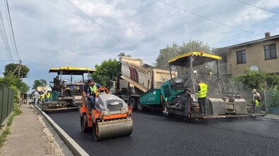 Процес асфальтування дороги, працює кілька одиниць техніки та робітники. На передньому плані видно покладений новий шар асфальту, який ущільнює помаранчевий каток. Також працюють два асфальтоукладачі та вантажівка, що вивантажує суміш.