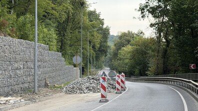 Ділянка дороги, де тривають роботи з укріплення схилу. Ліворуч видно нову підпірну стінку з габіонів і насип каміння, а місце робіт огороджене сигнальними конструкціями.