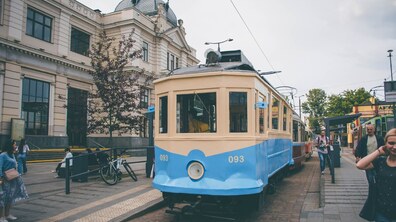 Біло-блакитний ретротрамвай стоїть на коліях біля Головного залізничного вокзалу. Навколо трамвая перебувають перехожі, а на другому плані видно історичну будівлю вокзалу. Фото з архіву ЛКП «Львівелектротранс», 2025 рік, автор - Володимир Караїм