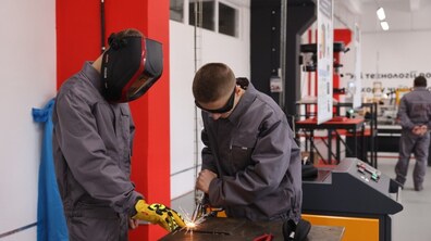 Двоє чоловіків працюють із металом. Для захисту обличчя вони використвують захисне спорядження. Один із них виконує обробку металу інструментом