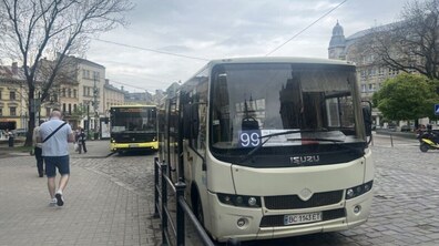 Білий автобус № 99 стоїть у центрі Львова, позаду нього – жовтий автобус. На тлі видно старовинну архітектуру міста, дерева та перехожих.