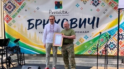 Перший заступник міського голови Львова Андрій Москаленко та керівник добровольчого формування Володимир Параняк стоять на сцені на тлі банера з написом «Брюховичі». Володимир Параняк тримає в руках подяку.