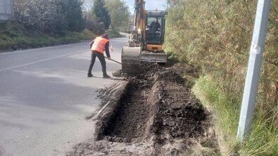 на узбіччі дороги невеликий екскаватор та робітник у жилеті проводять земляні роботи для облаштування тротуару