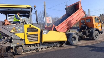 Процес укладання асфальту на дорозі за допомогою важкої будівельної техніки. Великий жовтий асфальтоукладач завантажується гарячою асфальтовою сумішшю з помаранчевої вантажівки-самоскида, що стоїть поруч. Позаду техніки видніється свіжовкладена чорна смуга асфальту